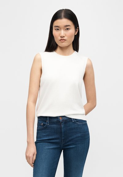 Γυναίκα με μακριά μαύρα μαλλιά, φοράει λευκό μπλουζάκι χωρίς μανίκια και μπλε τζιν, στέκεται μπροστά σε απλό ανοιχτό γκρι φόντο.