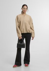 Béžová bomber bunda na zip s oversize střihem, černé zvonové džíny, červené a béžové platformové boty a černá kabelka se stříbrnými akcenty.