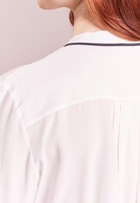 Blouse blanche à texture douce, avec une bordure contrastée foncée le long de l'encolure et un dos plissé.