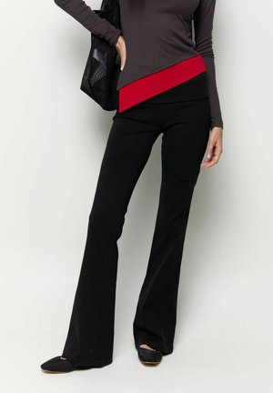 Vrouw draagt zwarte uitlopende broek, een donker topje met lange mouwen en een rode gelaagde zoom, zwarte platte schoenen, en draagt een zwarte tas over de schouder.
