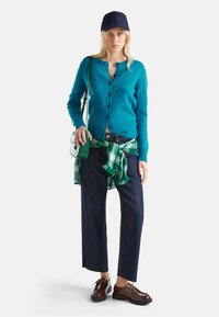 Donna che indossa un cardigan blu, una camicia a quadri verde legata in vita, jeans neri a gamba ampia, scarpe marroni, un berretto blu navy e una borsa a spalla verde su sfondo bianco.