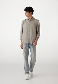 Camisa de botones gris claro con un bolsillo en el pecho, mangas enrolladas; combinada con jeans azul claro y zapatillas blancas. Ajuste simple y relajado.