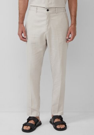 Mann trägt beige gerade geschnittene Hose mit vorderem Knopf und schwarze Sandalen mit Schnalle, steht vor einfachem Hintergrund.