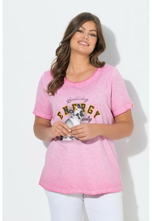 Donna con lunghi capelli castani che indossa una t-shirt rosa con un bulldog e la scritta "Solo energia", sorridente con le mani delicatamente intrecciate.