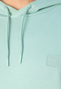 Mátově zelená mikina s kapucí, která má klokánčí kapsu, nastavitelná stahovací šňůrky s kovovými koncovkami a vyšívané logo "BOSS" na hrudi.