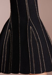 Détail de tissu noir côtelé avec des coutures dorées verticales formant des motifs triangulaires allongés sur une section évasée du vêtement.