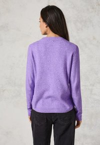 Lila Strickpullover mit langen Ärmeln und Rundhalsausschnitt, aus weichem Material; kombiniert mit schwarzen Cordhosen mit Gesäßtaschen.