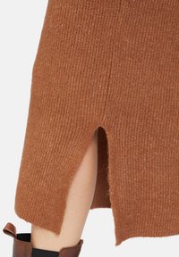 Stickad tröja i varmt brunt med ribbat mönster, med en sidoklyvning och en något trasig nederkant. Accentuerad med ett mjukt tygband.