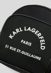 Zaino in nylon nero con testo bianco: "KARL LAGERFELD PARIS 21 RUE ST-GUILLAUME." Design arrotondato con dettagli lucidi.