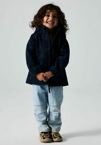 Veste navy avec capuche, texture à motifs, fermeture éclair à l'avant, associée à un pantalon en denim bleu clair et des baskets multicolores.