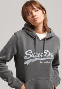 Hanorac gri cu şnur, buzunar frontal tip kangaroo și logo brodat "SuperDry Brooklyn". Material moale cu o croială lejeră și manșete ribbate.
