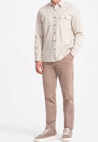 Camisa beige de manga larga con cierre de botones y dos bolsillos en el pecho, combinada con pantalones de pana marrón claro y zapatillas grises.