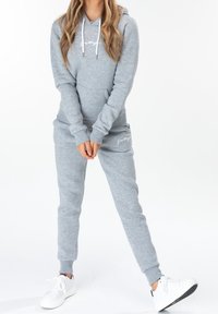 Personne portant un sweat à capuche gris clair et un jogging assorti avec des baskets blanches, debout les mains jointes et une jambe légèrement tendue.