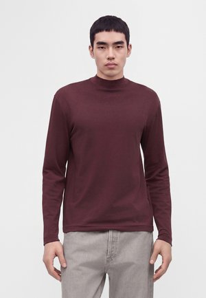 Jonge man draagt een bordeauxrode top met lange mouwen en opstaande kraag en lichtgrijze broek, staand tegen een effen witte achtergrond.
