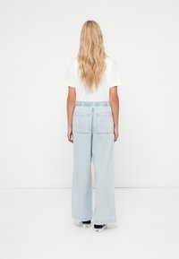 Világoskék, széles szárú farmer rugalmas derékpánttal és két hátul zsebbel, egyszerű fehér pólóval párosítva. A haj hullámos és hosszú.