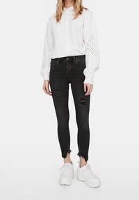 Camisa blanca tipo botón con detalles de ojales en las mangas, combinada con jeans negros ajustados que presentan áreas desgastadas. Llevada con zapatillas deportivas blancas de suela gruesa.