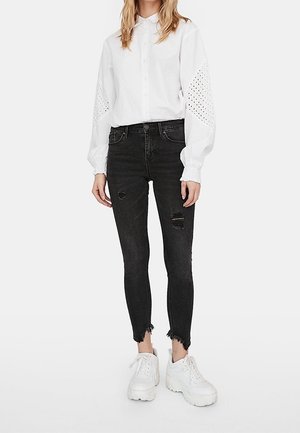 Weißes Hemd mit Knöpfen und Lochstickerei an den Ärmeln, kombiniert mit schwarzen Skinny Jeans mit Destroyed-Details. Getragen mit weißen, klobigen Turnschuhen.