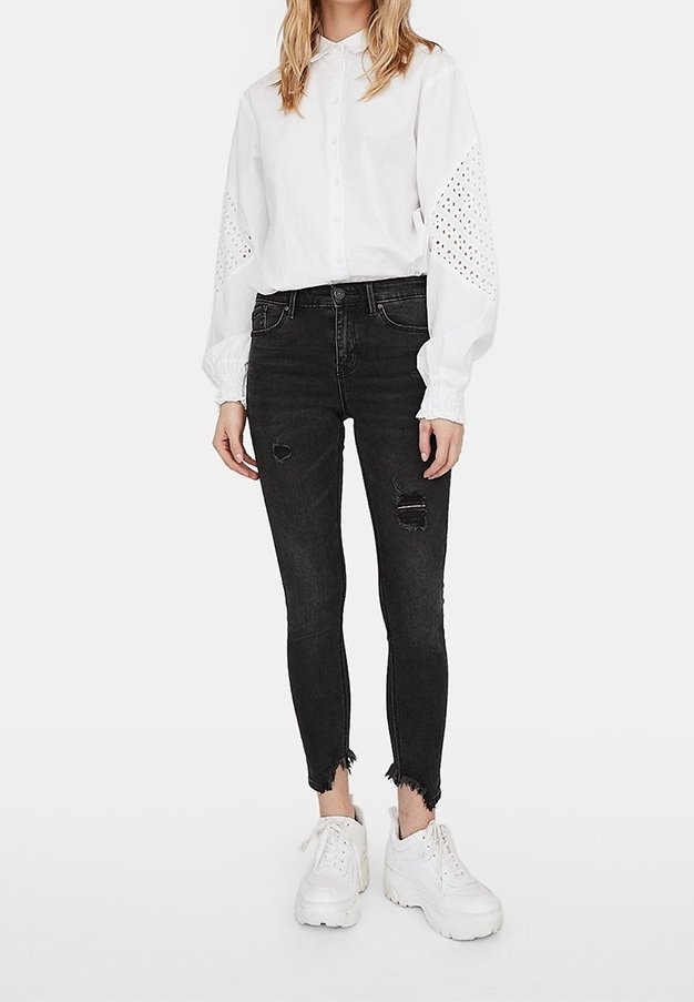 Camisa blanca tipo botón con detalles de ojales en las mangas, combinada con jeans negros ajustados que presentan áreas desgastadas. Llevada con zapatillas deportivas blancas de suela gruesa.