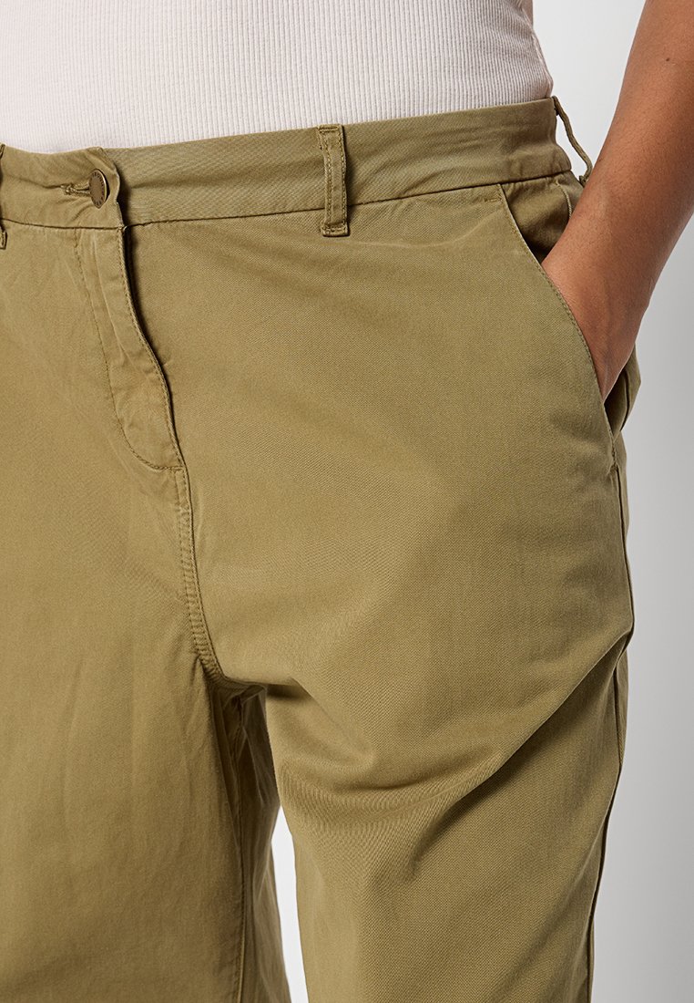 Nahaufnahme einer Person in khakifarbigen Hosen, mit einer Hand in der Seitentasche und einem beigefarbenen, gerippten Oberteil, das in die Hose gesteckt ist.