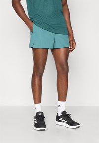 Shorts de sport pour hommes en teal, tissu léger avec poches latérales, associés à des chaussures de course noires et des chaussettes de sport blanches.
