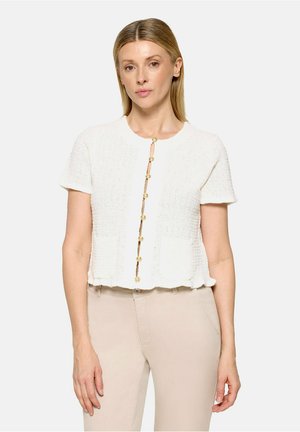 Kvinde iført en cremefarvet tekstureret kortærmet cardigan med guldknapper og beige højtaljede bukser mod en ensfarvet baggrund.