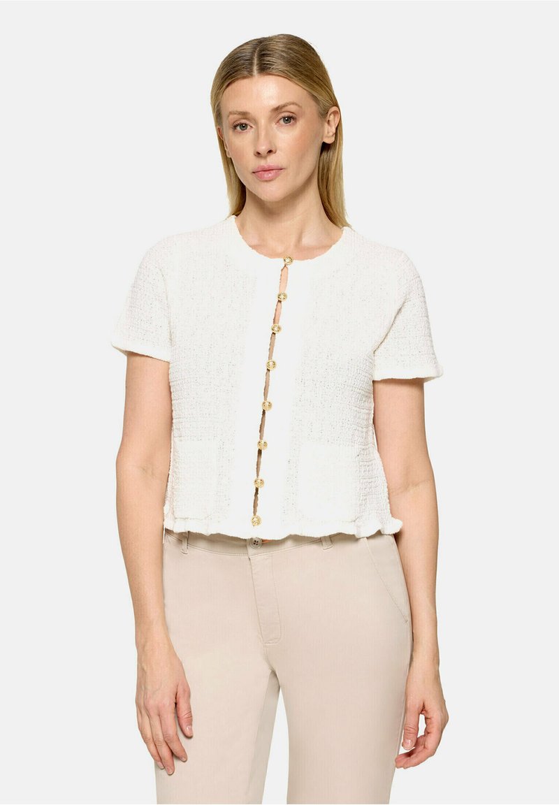 Femme portant un cardigan texturé crème à manches courtes avec des boutons dorés et un pantalon beige taille haute sur un fond uni.