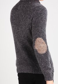 Grauer Strickpullover mit hohem Kragen und strukturiertem Körper. Auffällige offene Strick-Ärmelflicken sorgen für Kontrast. Weiches, warmes Material mit Sprenkeln.