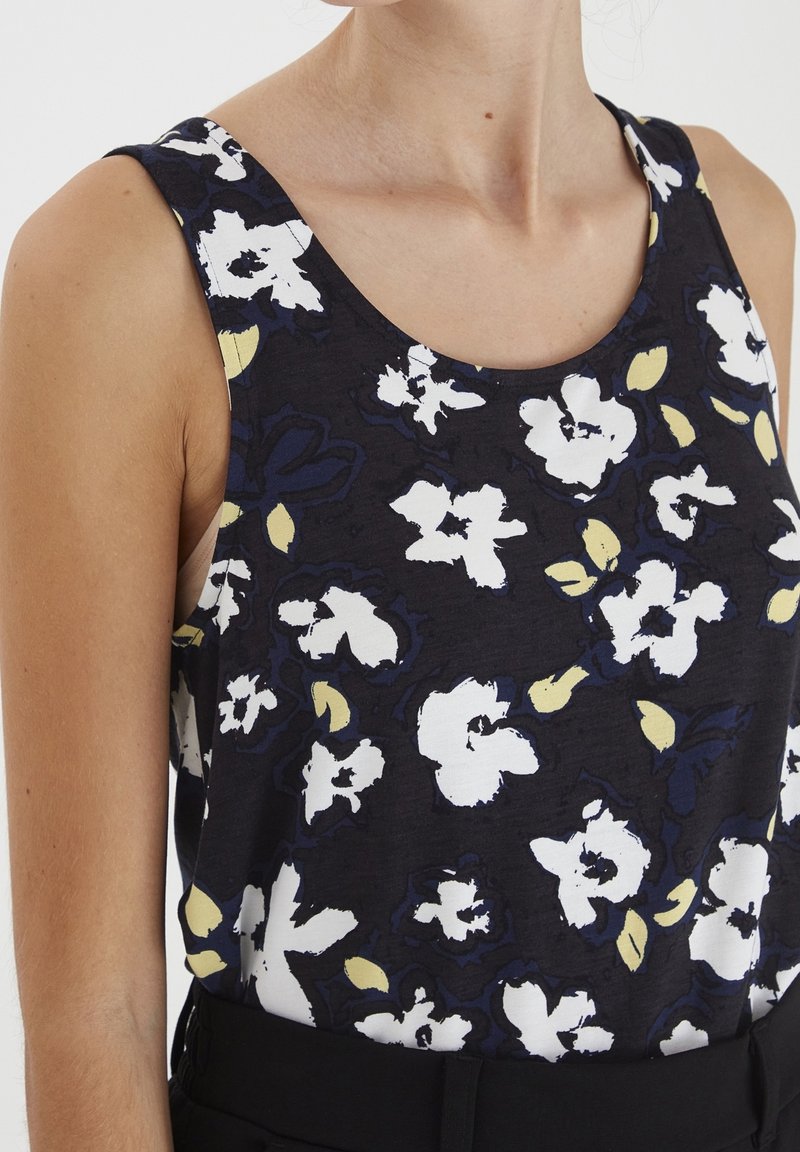 Top negro sin mangas con un patrón floral en blanco y amarillo, llevado metido dentro de pantalones negros, mostrando el torso superior y el cuello.