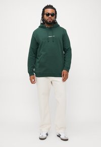 Hanorac verde închis cu buzunar frontal, șnur de reglare și detalii cu logo, asociat cu pantaloni crem și adidași alb-negru.