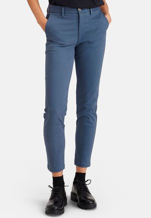 Osoba ubrana w obcisłe, niebieskie spodnie 7/8 z przednimi kieszeniami, czarne skarpety i czarne sznurowane buty, stojąca na jednolitym tle.