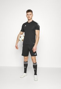Zwarte sporttrui en -shorts met witte strepen en logo, textuurstof, gecombineerd met zwarte sokken en witte voetbalschoenen, terwijl hij een bal vasthoudt.
