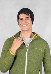 Groene, textuurgeweven zip-up jas met gele accenten, grijs t-shirt eronder en een marineblauwe gebreide beanie. Model glimlacht.