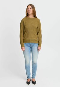 Olívazöld kötött pulóver texturált mintával, világoskék farmernadrággal és fekete hegyes orrú balerinacipővel kombinálva, laza fazonnal és kerek nyakkivágással.