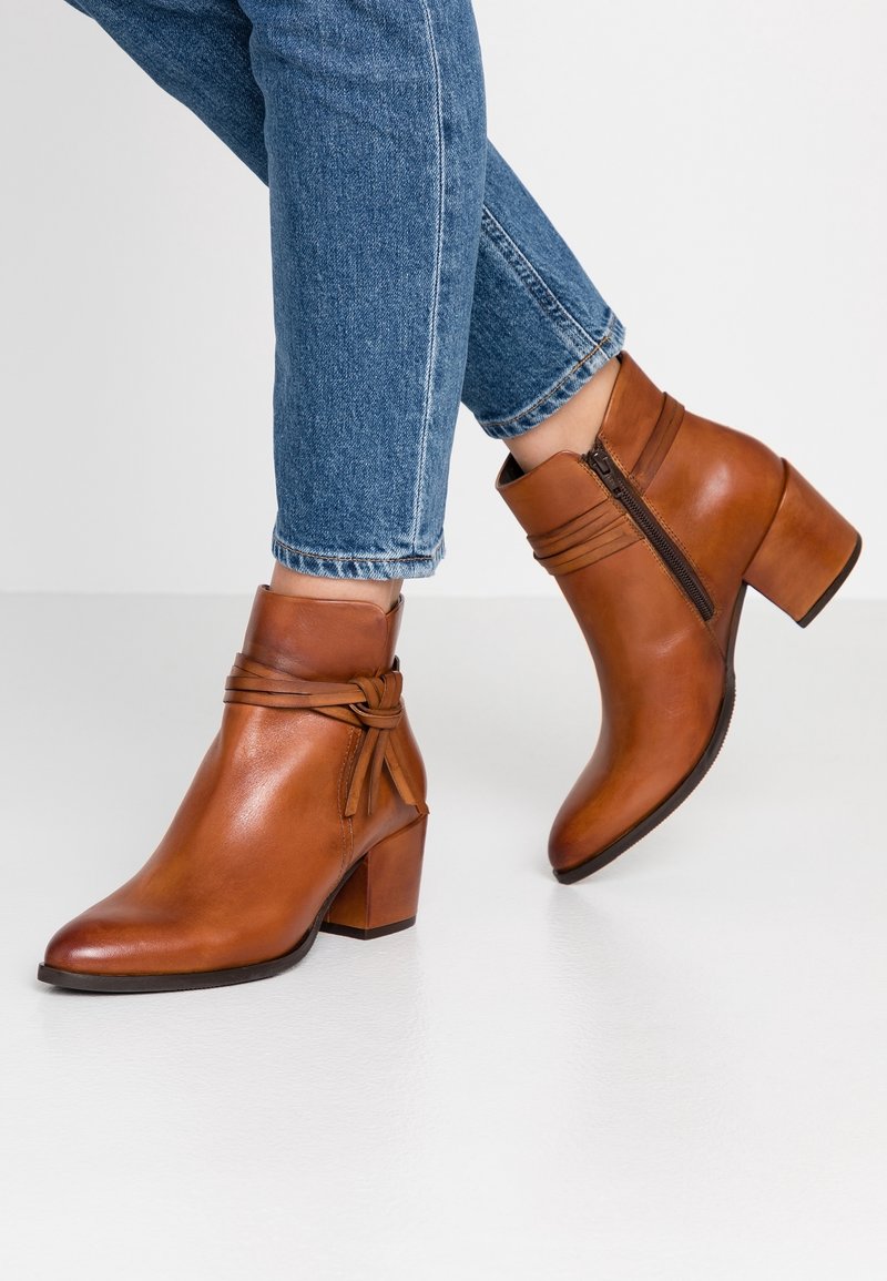 Femme portant un jean bleu et des bottines en cuir marron avec une fermeture éclair sur le côté et un détail de nœud décoratif, debout sur un sol blanc.