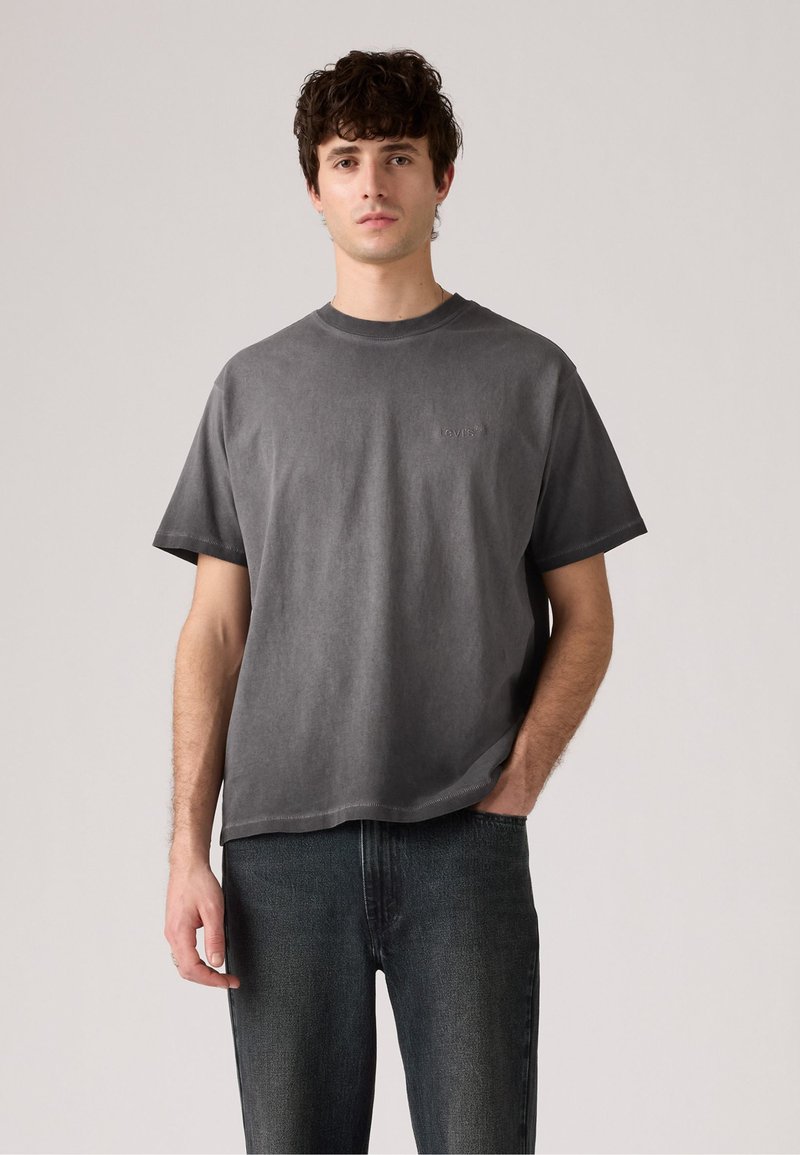 T-shirt en coton gris foncé avec un col rond et des manches courtes, arborant un logo subtil brodé sur la poitrine. Associé à un jean foncé.