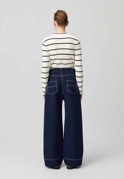 Pantalons larges bleu marine avec des poches arrière bien visibles, associés à un pull crème à rayures horizontales noires et un col festonné.