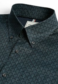 Dunkelgrünes gemustertes Hemd mit Button-Down-Kragen, das ein fächerartiges Design aufweist, schwarze Knöpfe und ein navyblaues Innenfutter mit Punkten.