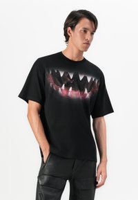 Young man wearing black T-shirt with large graphic of open shark mouth showing sharp teeth, paired with black cargo pants.