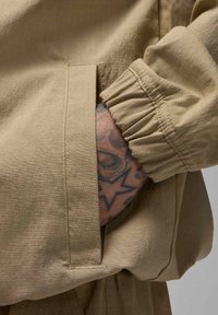 Țesătură de jachetă bej cu finisaj texturat, dotată cu un buzunar lateral și manșete pliate. Tatuaj expus vizibil lângă mânecă.