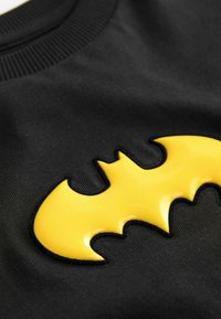 Čierna látková mikina s lesklým žltým logom Batmana. Dizajn zvýrazňuje hladkú textúru s ryhľovým detailom na krku.