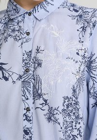 Sininen raidallinen paita, jossa on merkkikukkakuvioita tummansinisellä ja valkoisella, nappikoristeinen malli ja terävä kaulus. Kangas näyttää kevyeltä.