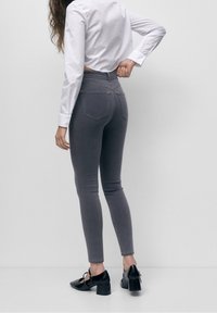 Femme portant un jean skinny taille haute gris, une chemise blanche à manches longues et des chaussures Mary Jane à talons blocs noirs, debout devant un fond uni.