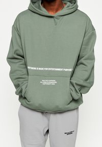 Olivgrüner Kapuzenpullover mit Kängurutasche, mit weißem Textdesign auf der Vorderseite und einer lockeren Passform. Kombiniert mit grauen Jogginghosen.