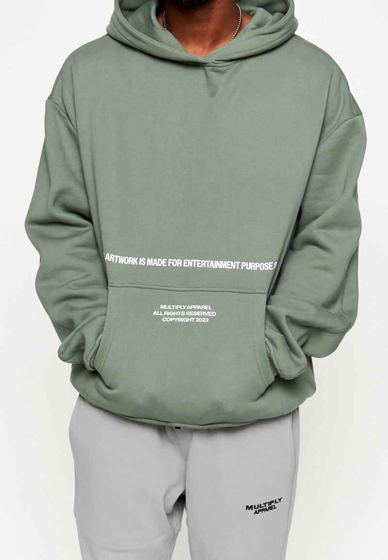 Olivgrüner Kapuzenpullover mit Kängurutasche, mit weißem Textdesign auf der Vorderseite und einer lockeren Passform. Kombiniert mit grauen Jogginghosen.