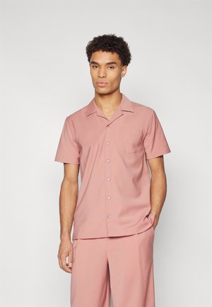 Chemise à manches courtes, boutonnée, en tissu rose clair avec une poche, col en V et texture lisse ; portée avec un pantalon assorti.