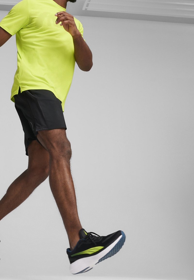 Homme portant un T-shirt jaune néon, un short noir et des chaussures de course noires marchant devant un fond gris uni.