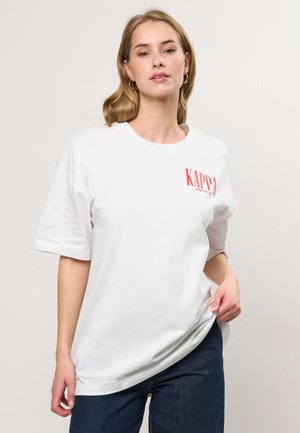 Mujer rubia con pendientes de aro dorados, camiseta blanca de gran tamaño de Kappa con logo rojo y jeans azul oscuro, de pie contra un fondo claro.