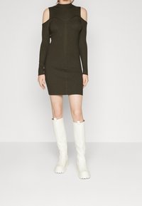 Olivgrünes, geripptes Strickkleid mit Cut-Outs an den Schultern, langen Ärmeln und figurbetontem Design; kombiniert mit weißen kniehohen Stiefeln.