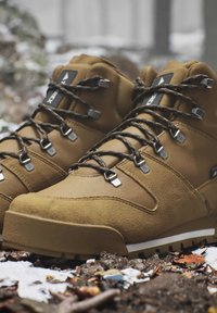Brown hiking boots with black and yellow laces on snowy and rocky ground outdoors.