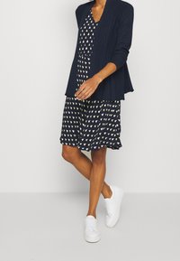 Giacca a coste navy sopra un abito coordinato con motivi triangolari verdi e bianchi. Completato con sneaker bianche, evidenziando un look stratificato.
